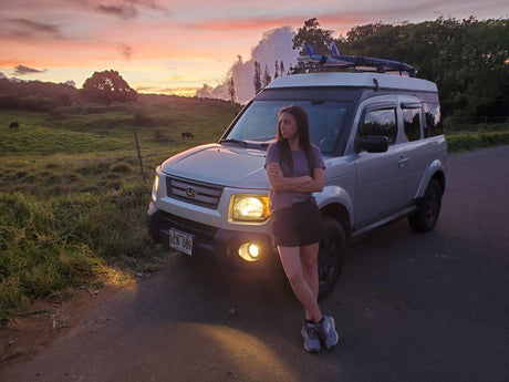 Sunset adventure: A woman by her Honda Element RV rental with pop-top roof and surfboards. Headlights glow by a grassy field.