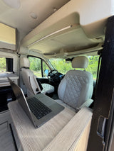 Rv rental interior: a comfy mobile office with light-colored, quilted seats, a laptop on a wood-grain desk, and a green view outside.