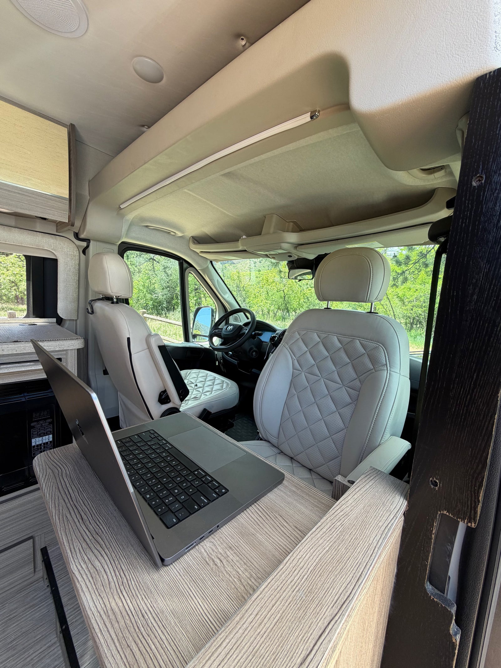 Rv rental interior: a comfy mobile office with light-colored, quilted seats, a laptop on a wood-grain desk, and a green view outside.