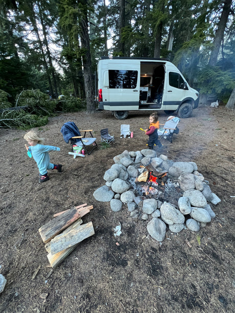 An RV rental is parked at a wooded campsite, where two young children play near a large, stone-ringed campfire.
