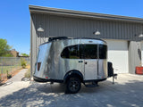 A silver RV rental is parked outside a gray metal building under a clear blue sky, ready for outdoor adventures.