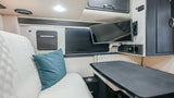 Spacious rv rental interior showing white quilted seating, a large table, and a mounted TV, ideal for comfortable travel or work.