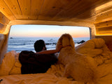 From a comfortable RV rental bed, a man and his dog gaze at a serene ocean sunset.