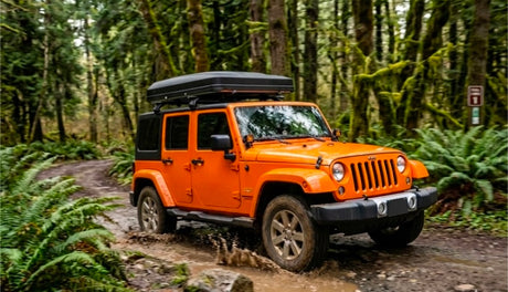 An orange Jeep Wrangler with a roof-top tent navigates a muddy forest trail. Explore nature with an RV rental!