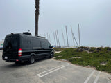 A sleek black Mercedes Sprinter RV rental parked on a paved lot by a sandy beach with sailboats and a misty sky.