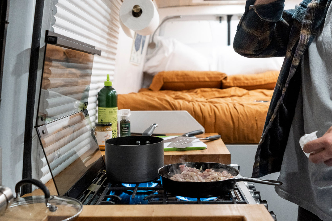 A person cooks in an RV rental kitchen, with meat sizzling on the gas stove and a cozy bed in the background.