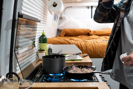 A person cooks in an RV rental kitchen, with meat sizzling on the gas stove and a cozy bed in the background.