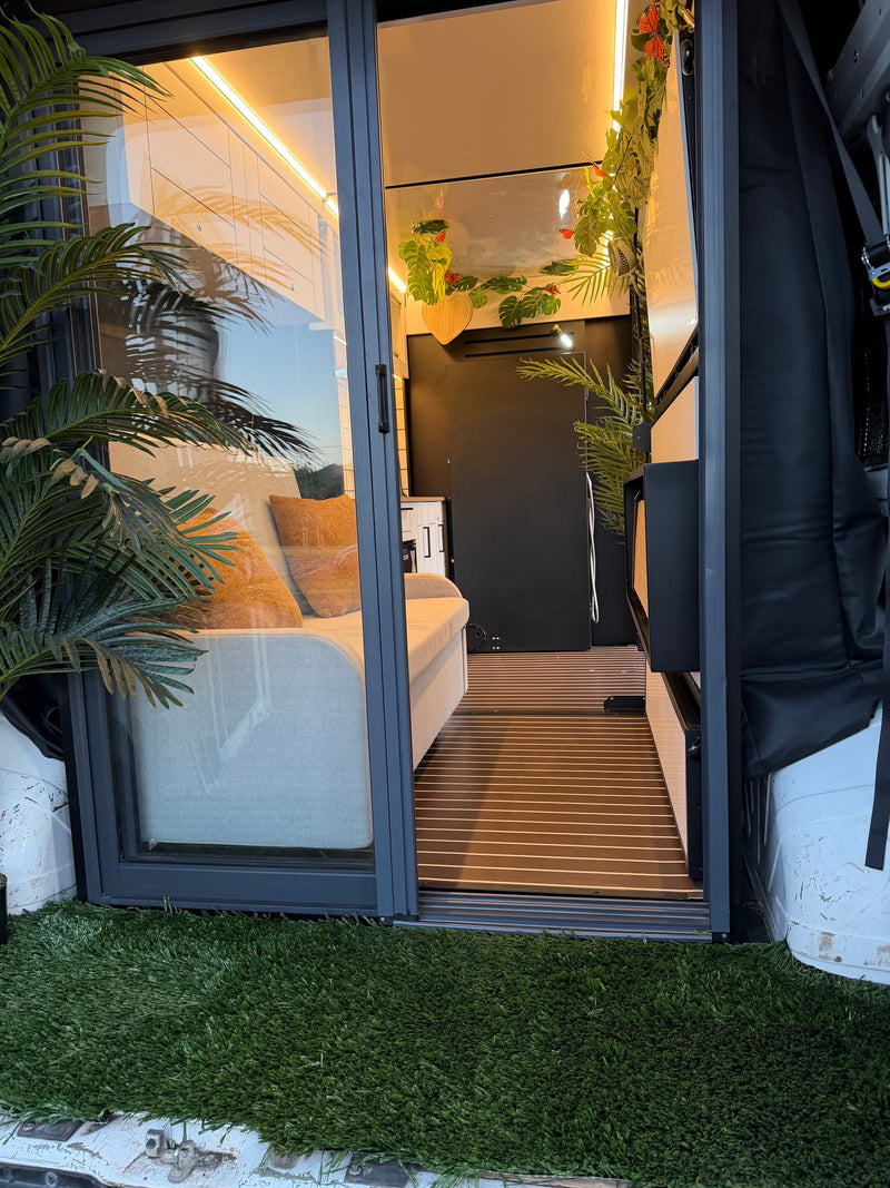 View of a modern rv rental's cozy camper van interior through a glass door, featuring warm lighting, green plants, and a comfortable couch. — photo 3