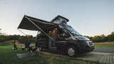 This RV rental camper van is set up for a relaxing campsite adventure, with an awning extended & pop-top raised.