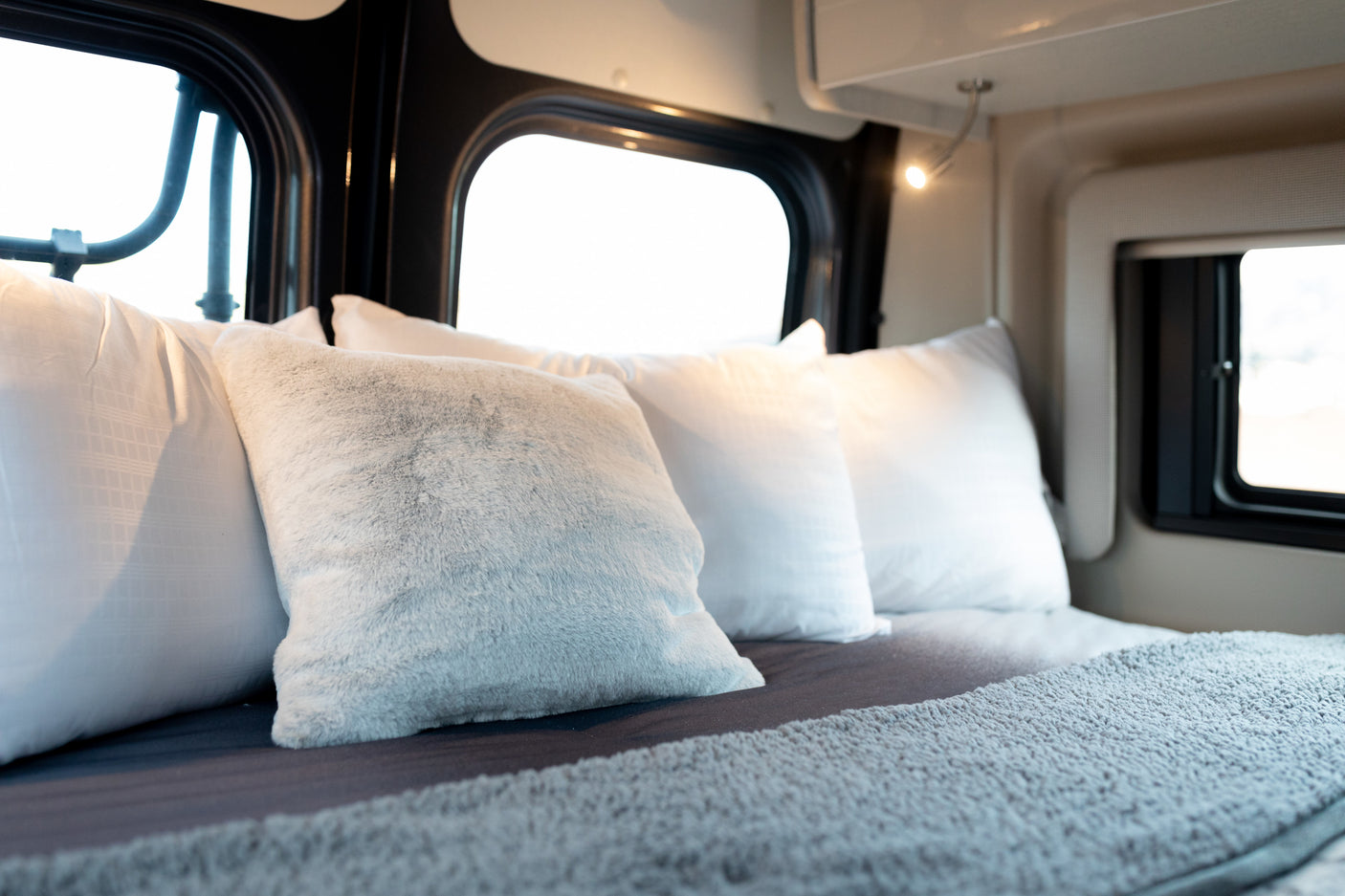 Cozy RV rental bedroom with white and grey pillows, a soft blanket, and large bright windows for a comfortable travel interior.