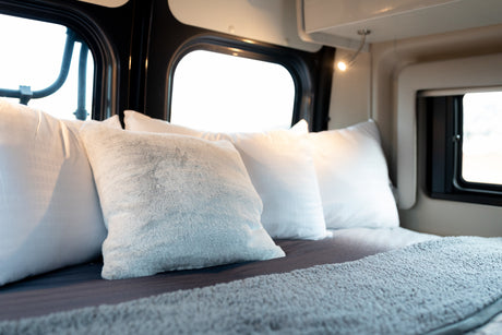 Cozy RV rental bedroom with white and grey pillows, a soft blanket, and large bright windows for a comfortable travel interior.