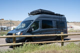 This dark gray Ford Transit camper van RV rental with rooftop tent and ladder is parked outdoors by a wooden fence.
