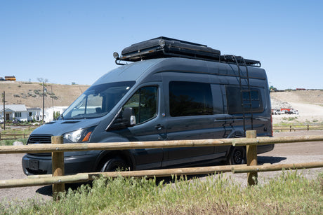 This dark gray Ford Transit camper van RV rental with rooftop tent and ladder is parked outdoors by a wooden fence.