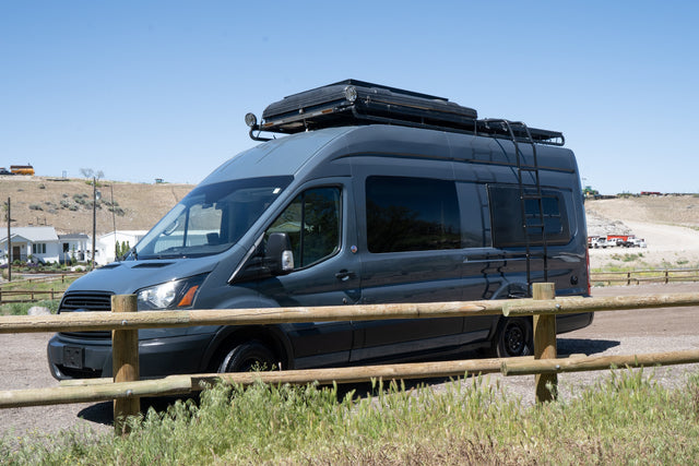 This dark gray Ford Transit camper van RV rental with rooftop tent and ladder is parked outdoors by a wooden fence.