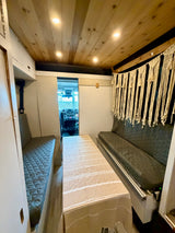 View inside an RV rental with a wooden ceiling, recessed lights, two quilted benches, a central table, and macrame wall art.