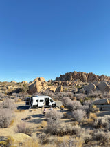 An **rv rental** parked among massive rock formations in a sunny desert campsite, with two people relaxing at a picnic table.