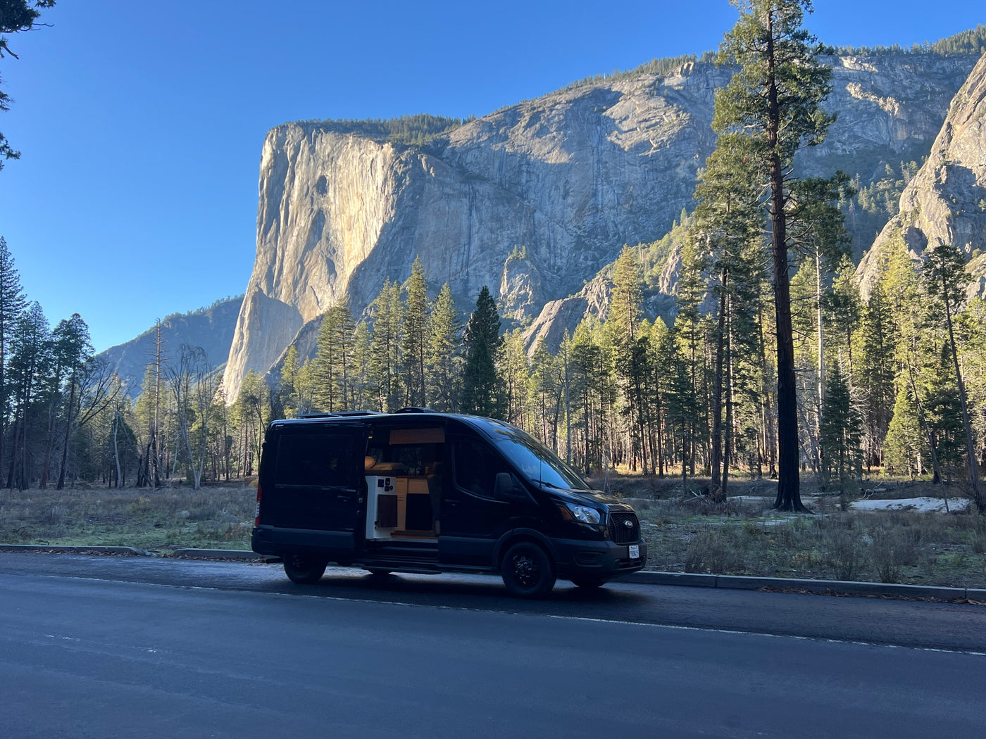 An RV rental van with its side door open is parked by a towering granite cliff and lush forest on a bright, clear day. — photo 19