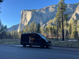 An RV rental van with its side door open is parked by a towering granite cliff and lush forest on a bright, clear day.