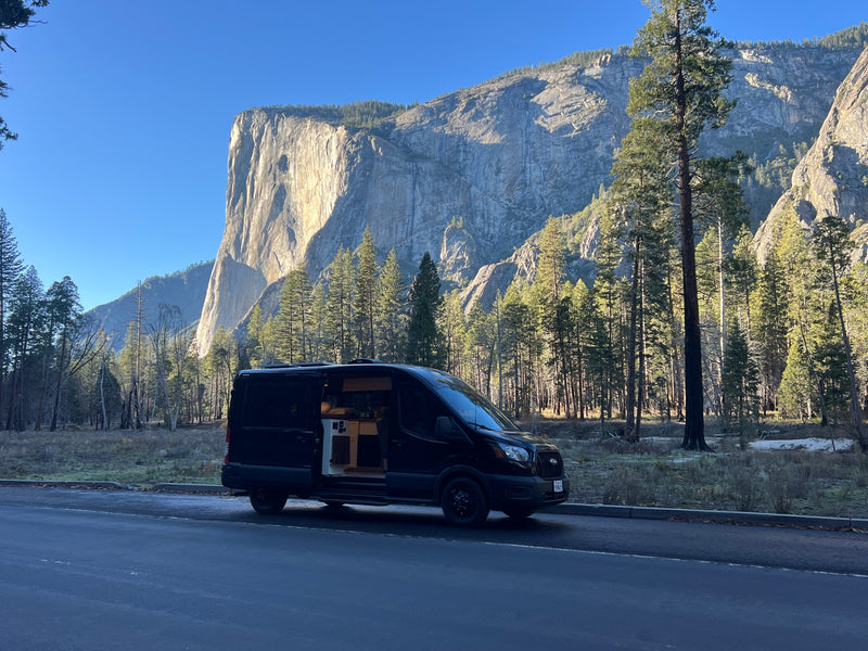 An RV rental van with its side door open is parked by a towering granite cliff and lush forest on a bright, clear day. — photo 19