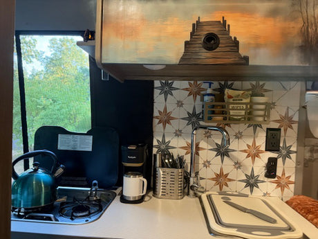 Wanda's galley - Upper cabinet with artwork holds dishes, decorative backsplash, sink, coffee pot, utensils, and propane stove top with tea kettle.