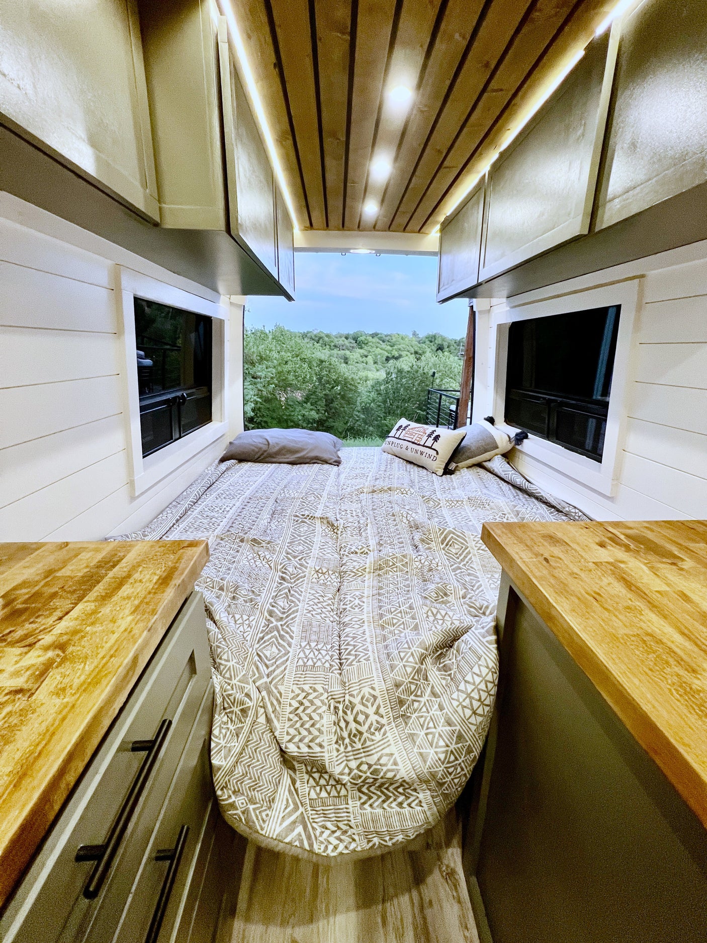 Cozy camper van RV rental interior: bed with patterned bedding, wood ceiling, and panoramic forest views through open doors.