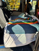 Cozy RV rental van interior with bed, colorful blanket, pillows, and an autumn tree view through the window.