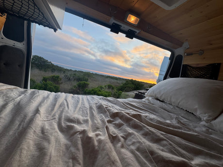 Relaxing in an RV rental campervan bed with a beautiful sunset view over a natural landscape and campsite.