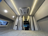 Cozy RV rental interior: bed in foreground, bright ceiling lights, AC unit, and front cabin view for travel comfort.