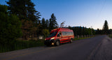 A bright red RV rental, a camper van, illuminates a scenic road at twilight, nestled among trees and mountains.