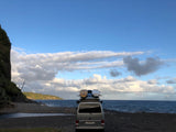 This rv rental camper van with surfboards enjoys a scenic ocean and beach view, nestled by a green cliff under a cloudy sky.