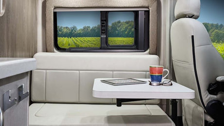 Inside a bright RV rental: a compact living space with table, tablet, mug, and glasses. Enjoy vineyard views through the window.