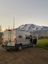Silver RV rental on a dirt road, facing a snow-capped mountain backdrop with bare trees. Ideal for outdoor adventure.