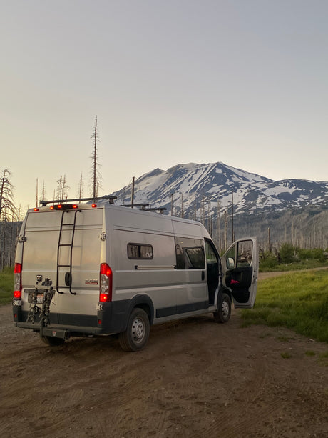 Silver RV rental on a dirt road, facing a snow-capped mountain backdrop with bare trees. Ideal for outdoor adventure.