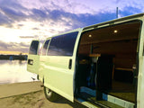 White camper van, side door open to its custom interior, parked at water's edge under a colorful sunset sky. Ideal RV rental.