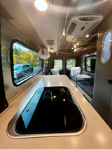 Inside an RV rental, a sleek kitchen counter and cooktop lead to comfy seating and large windows with outdoor views.
