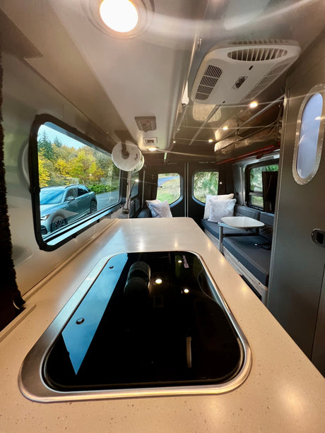 Inside an RV rental, a sleek kitchen counter and cooktop lead to comfy seating and large windows with outdoor views.