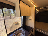 Inside a modern rv rental: kitchenette with sink & induction cooktop, overhead cabinet, and bed. Screened window shows a pine forest view.