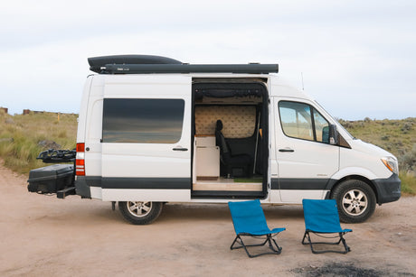 White RV rental Sprinter camper van with open door and interior view. Two blue chairs await a camping adventure.