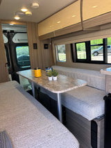 RV rental interior featuring a cozy dining area with benches, a table, mugs, and plants, offering a scenic window view.