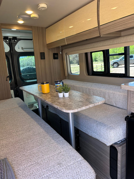 RV rental interior featuring a cozy dining area with benches, a table, mugs, and plants, offering a scenic window view.