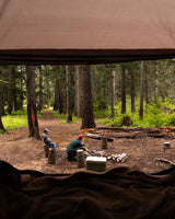 View from an RV rental showing two people camping in a forest, preparing a campfire by log seats and a cooler.