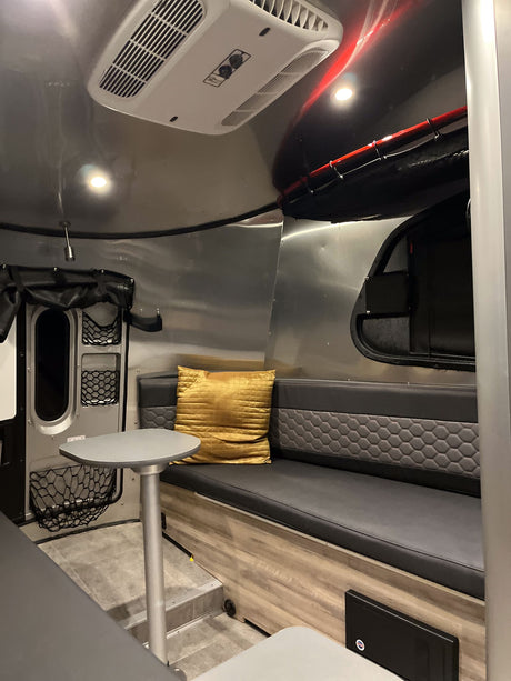 RV rental interior: A modern living space with curved aluminum walls, a grey bench seat, golden pillow, and overhead AC unit.