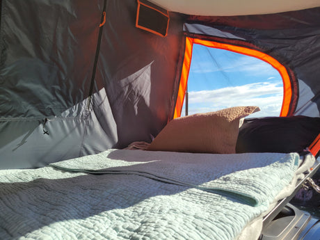 Relax in this RV rental's cozy rooftop tent, featuring a comfy bed with a light blue blanket and a clear sky view through the orange-trimmed opening.