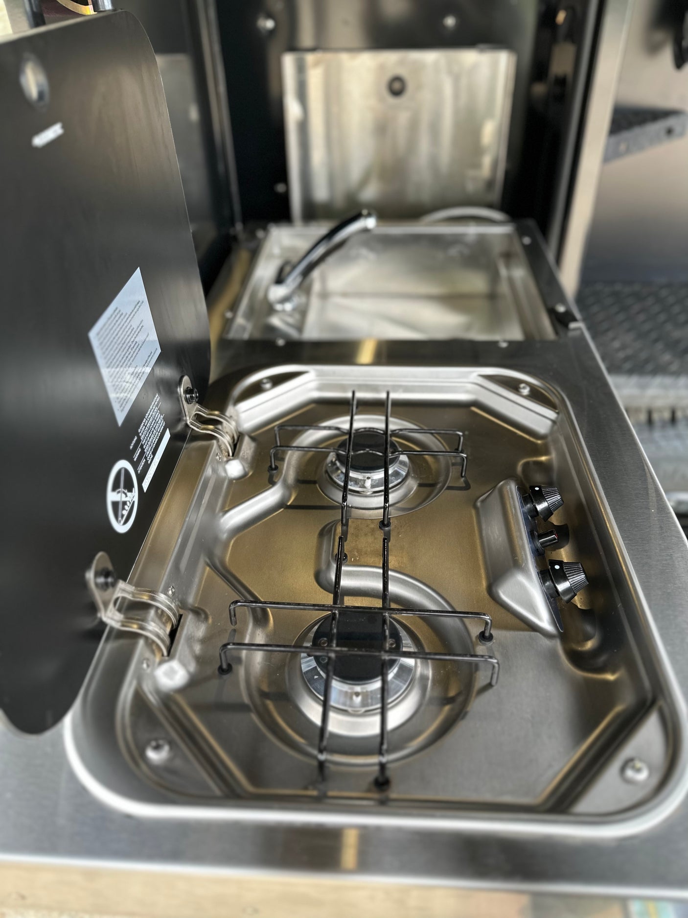 Compact stainless steel RV kitchen with dual gas burners, sink, and safety-labeled cover, ideal for rv rental adventures.