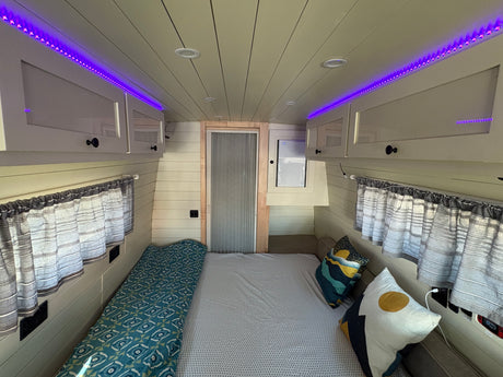 Cozy RV rental interior with a comfortable bed, vibrant purple LED lighting, overhead cabinets, and window curtains.