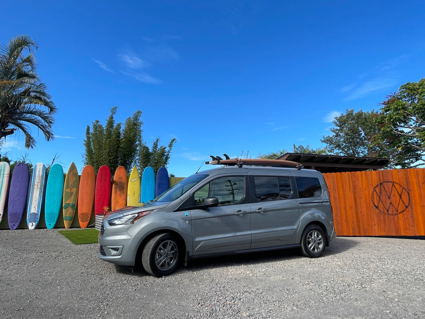 Gray van with surfboard on roof, parked by colorful surfboards. This could be your next RV rental adventure!