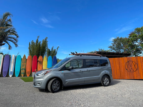 Gray van with surfboard on roof, parked by colorful surfboards. This could be your next RV rental adventure!