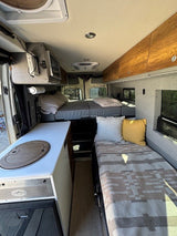 Inside an RV rental: a cozy camper van interior with a galley, elevated rear bed, and side bench/bed.