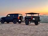 A dark Jeep tows an off-road utility trailer at sunset, overlooking a city. Ideal setup for an RV rental adventure.