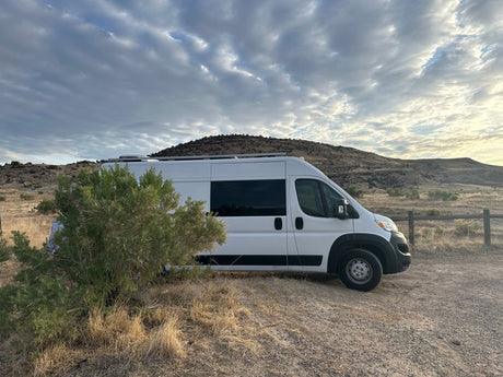 Scenic RV rental: White camper van parked on a dirt road, surrounded by hills and a cloudy, expansive sky.